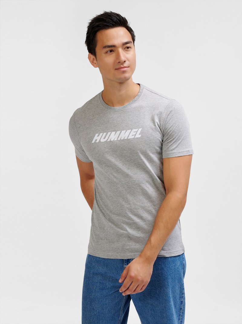 Joven de pie, vistiendo una camiseta gris con el logo de "HUMMEL" y vaqueros azules, mirando ligeramente hacia un lado contra un fondo liso.