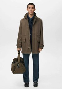 Chaqueta marrón de botones con bolsillos, llevada sobre una blusa oscura, combinada con jeans oscuros y zapatos negros; sostiene una mochila de viaje verde.