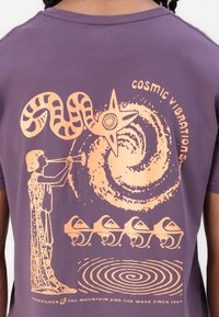 Purple cotton t-shirt featuring orange graphic design, depicting a musician with a trumpet, a swirl, and abstract shapes with mountain accents.