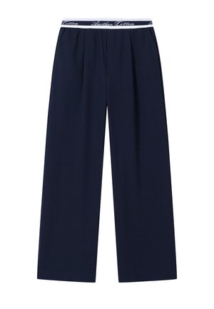 Tengerészkék bőszárú nadrág redőkkel és fehér elasztikus derékszalaggal, amelyen dőlt betűkkel az "Another Cotton" felirat olvasható.