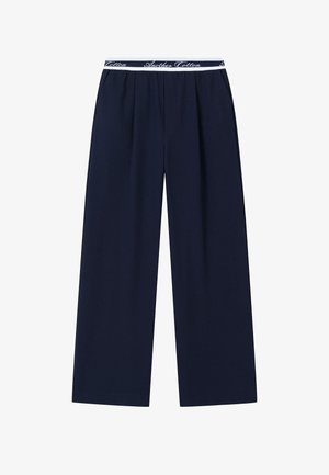 Tengerészkék bőszárú nadrág redőkkel és fehér elasztikus derékszalaggal, amelyen dőlt betűkkel az "Another Cotton" felirat olvasható.