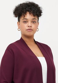 Woman with curly hair wearing gold hoop earrings and a burgundy cardigan over a white top, facing forward with a neutral expression.