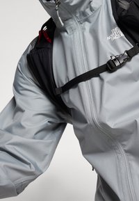 Vaaleanharmaa vedenpitävä takki, jossa korkea kaulus, vetoketjusuljenta ja säädettävät hihnat. Takissa on brändäys ja mustat kontrastielementit.