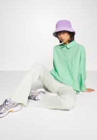 Camisa oversized verde-claro com botões, calças verde-claro caneladas, chapéu de balde roxo e ténis cinzentos com detalhes laranja e roxo.