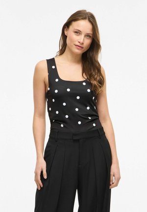 Kobieta w czarnym topie bez rękawów w białe kropki i czarnych plisowanych spodniach z wysokim stanem na białym tle.