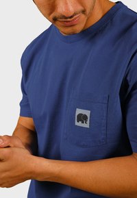 Mörkblå bomullströja med rund hals och korta ärmar. Har en grå ficka med en svart elefantlogotyp. Slät textur.