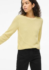 Πλεκτό κίτρινο πουλόβερ με υφή, λαιμόκοψη στρογγυλή, μακριά μανίκια και άνετη εφαρμογή. Συνδυασμένο με μαύρο παντελόνι.