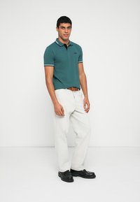 Kékeszöld galléros póló csíkos gallérral, világos színű nadrággal és fekete cipővel kombinálva. A pólón mellrészen hímzett logó látható.