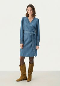 Robe en denim avec des manches longues, une taille nouée et un détail de poignet smocké. Associée à des bottes en suède marron jusqu'aux genoux.