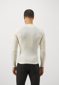 Langärmliges, hellbeiges Thermoshirt mit taillierter Passform, glatter Textur und flachen Nähten. Kombiniert mit enganliegenden schwarzen Sportleggings.