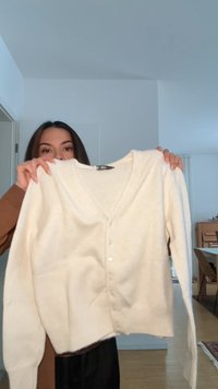 Mujer sosteniendo un cárdigan blanco abotonado en una habitación bien iluminada con suelo de madera y muebles al fondo.