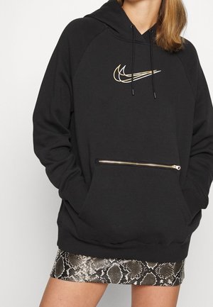 Vrouw draagt een zwarte hoodie met een gouden dubbele swoosh-logo en een voorzak met rits, gecombineerd met een rok met slangenhuidprint.