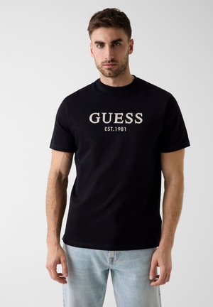 Uomo con capelli corti castani che indossa una T-shirt nera Guess con la scritta "EST. 1981", abbinata a jeans azzurri chiari, in piedi davanti a uno sfondo semplice.