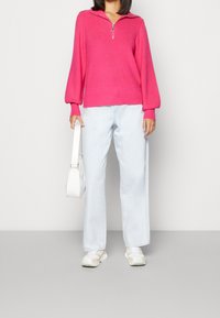 Mujer vestida con un suéter de manga larga de color rosa brillante con cremallera, jeans de pierna ancha de color azul claro, zapatillas blancas y sosteniendo un bolso blanco.