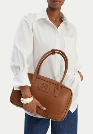 Persona con camicia bianca e jeans blu tiene una borsa in pelle marrone con logo "GR" impresso e un bracciale d'oro al polso.