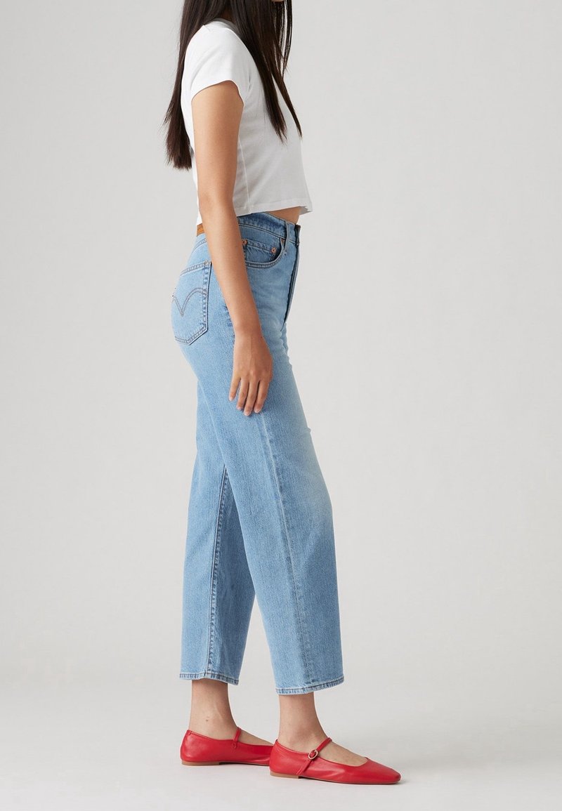 Femme en jean taille haute bleu clair, t-shirt court blanc et ballerines rouges Mary Jane, debout sur un fond gris.
