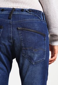 Jeans de mezclilla oscura con una textura suave. Presentan dos bolsillos traseros; uno con un acento en negro contrastante y un detalle de costura minimalista.