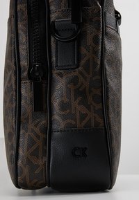 Sac à bandoulière marron à motifs présentant un design texturé avec des accents en cuir noir et une fermeture éclair visible sur un côté. Logo estampillé à la base.