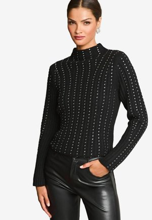 Schwarzer gestrickter Pullover mit hohem Kragen, der vertikales Rippenmuster und metallische Nieten aufweist. Getragen mit enganliegenden schwarzen Lederhosen.