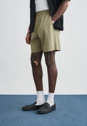 Khaki rövidnadrág elasztikus derékpánttal, oldalzsebekkel és bő szabással, fekete bebújós cipővel és fehér boka zoknival párosítva.