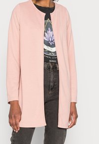 Kvinna som bär en ljusrosa öppen fram långärmad jacka över en svart grafik-t-shirt och mörkgrå jeans mot en vit bakgrund.