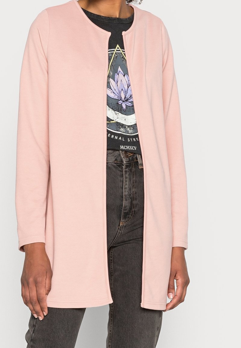 Kvinna som bär en ljusrosa öppen fram långärmad jacka över en svart grafik-t-shirt och mörkgrå jeans mot en vit bakgrund.