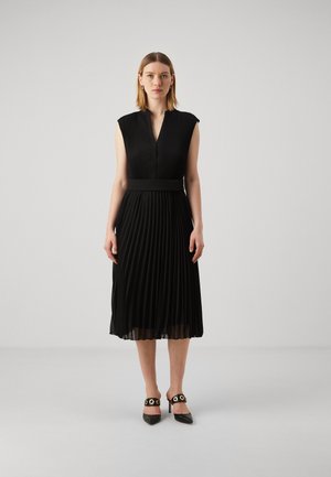 Black pleated dress with a v-neck, cap sleeves, and a cinched waist. Worn with pointed-toe heels featuring gold accents.