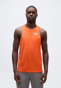 Νεαρός άνδρας που φοράει πορτοκαλί αθλητική φανέλα χωρίς μανίκια The North Face και γκρι παντελόνι, στέκεται μπροστά σε έναν απλό λευκό φόντο.