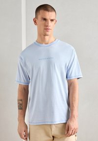 Světle modré bavlněné tričko s krátkým rukávem, kulatým výstřihem a jemně tištěným logem "SCOTCH & SODA" uprostřed. Hladká textura.
