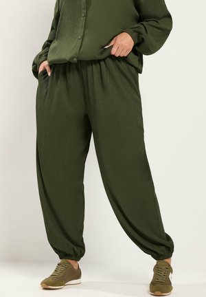 Persoon die losse olijfgroene, gestructureerde broek en bijpassend overhemd met elastische manchetten draagt, handen in de zakken, en olijfgroene sneakers met witte zolen.