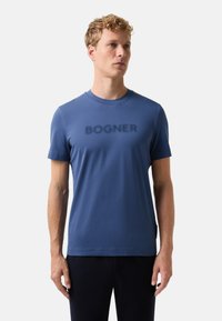 En blond mand iført en blå kortærmet T-shirt med "BOGNER" trykt på brystet, stående mod en ensfarvet lys baggrund.