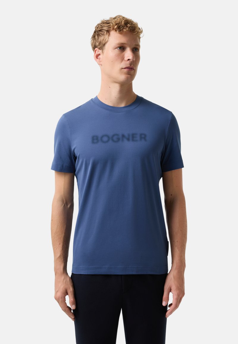 En blond mand iført en blå kortærmet T-shirt med "BOGNER" trykt på brystet, stående mod en ensfarvet lys baggrund.