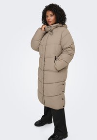 Beige lång vadderad jacka med huva, med sydda mönster, sidfickor och knappdetaljer. Mjuk textur, ledig passform.