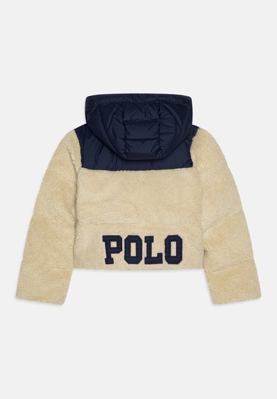 Rövidített dzseki, bézs műszőrme, tengerészkék steppelt kapucni és felső panel, tengerészkék "POLO" felirat a hátsó részen, puha tapintás, laza dizájn.