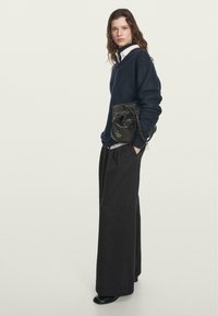 Marineblauwe trui met een relaxte pasvorm, gecombineerd met brede antracietkleurige broeken. Model houdt een kleine, gestructureerde zwarte tas vast. Eenvoudig, modern ontwerp.