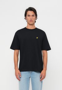 Camiseta de algodón negra con cuello redondo y mangas cortas, con un pequeño logo de águila amarilla en el pecho. Llevada con jeans de mezclilla azul claro.