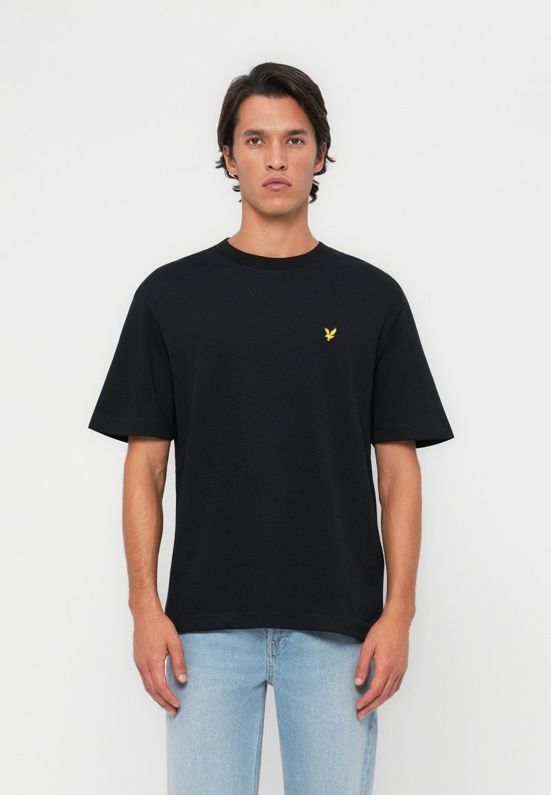 Camiseta de algodón negra con cuello redondo y mangas cortas, con un pequeño logo de águila amarilla en el pecho. Llevada con jeans de mezclilla azul claro.