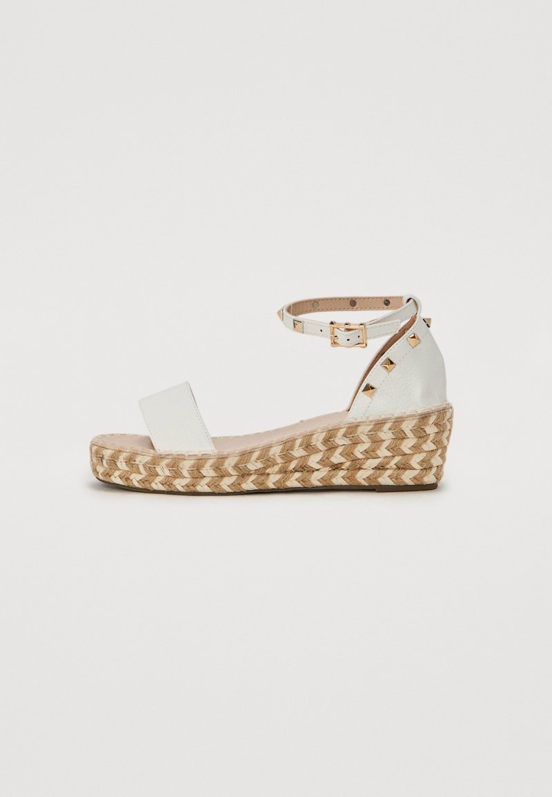 Sandales blanches à talons compensés avec plateforme en jute tressé, bride de cheville avec boucle dorée et clous pyramides dorés sur le contrefort.