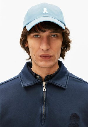 Jonge man met middellang bruin haar, die een lichtblauwe pet met een wit logo en een marineblauwe jas met rits draagt.