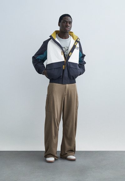 Νέος άνδρας στέκεται με τα χέρια στις τσέπες, φορώντας μια μπουφάν με μπλοκ χρωμάτων, γραφικό μπλουζάκι, φαρδιά καφέ παντελόνια και λευκά αθλητικά παπούτσια.