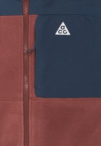 Giacca con zip rossa e blu navy dalla texture in pile, con un logo triangolare sulla tasca del petto. Sono visibili i dettagli delle cuciture.