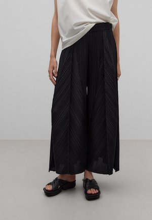 Persona con pantalones negros de pierna ancha plisados, camiseta blanca metida por dentro y sandalias negras de tacón con punta abierta, de pie sobre un suelo liso.