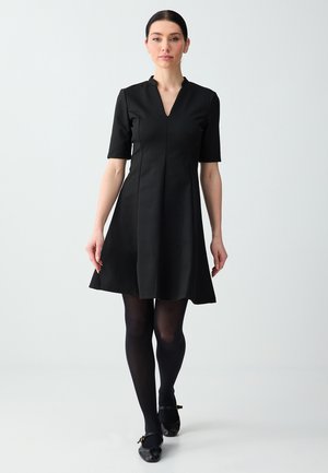 Kobieta o ciemnych włosach, ubrana w czarną sukienkę do kolan, czarne rajstopy i czarne buty, idąca do przodu na jasnym, jednolitym tle.