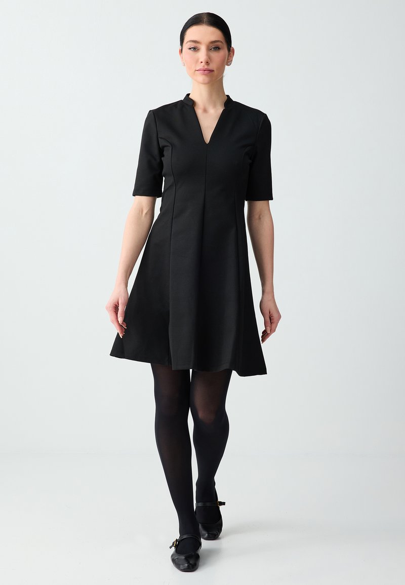 Femme aux cheveux foncés portant une robe noire jusqu'aux genoux, des collants noirs et des chaussures noires, marchant vers l'avant sur un fond clair uni.