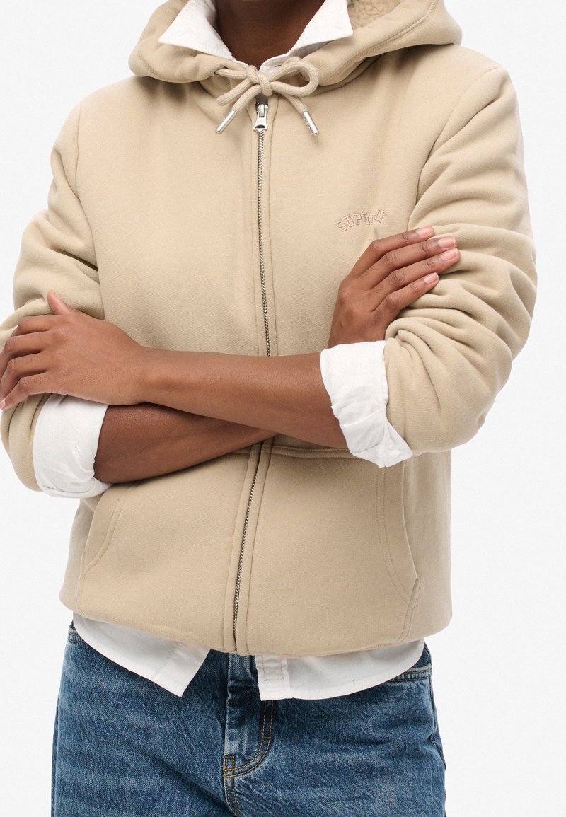 Haut à capuche zippé beige avec un cordon de serrage et un logo brodé. Présente une texture douce et des poignets côtelés sur une chemise blanche.