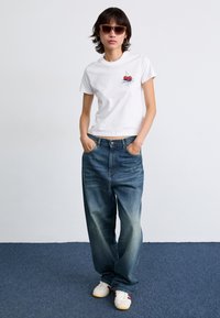 Camiseta blanca de algodón con gráfico de cereza, combinada con unos vaqueros de denim azul holgados. El modelo lleva gafas de sol marrones y zapatillas blancas con detalles rojos.
