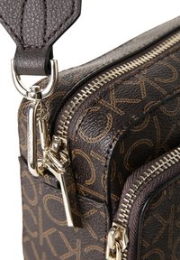 Brązowa torba ze skóry z fakturą, wyposażona w zamki błyskawiczne w złotym odcieniu oraz nadruk z logo. Zawiera odpinany pasek z srebrnym klipsem. Kompaktowy design.