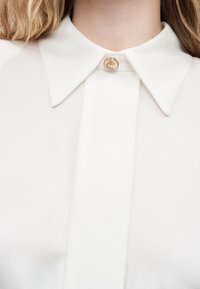 Λευκή μπλούζα με μυτερό γιακά κλειστό με ένα μόνο χρυσό κουμπί που διαθέτει υψωμένο λουλουδάτο σχέδιο.