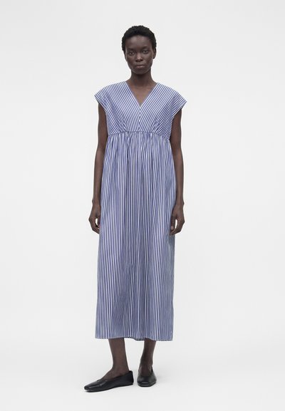 Γυναίκα φοράει ένα μπλε και λευκό κάθετο ριγέ αμάνικο μίντι φόρεμα με V-λαιμόκοψη και μαύρα ίσια παπούτσια, στέκεται μπροστά σε απλό φόντο.