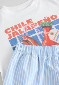 Bijela majica s grafikom "Chile Jalapeño", koja prikazuje crvene čili papričice i zelenu bočicu. Prikazana svijetloplava suknja s bijelim prugama.